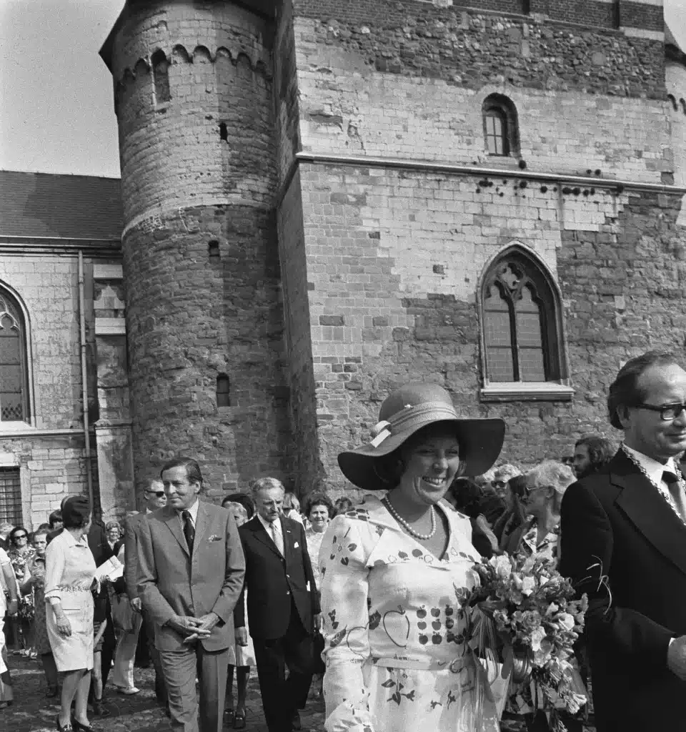 Black-and-white photograph: Princess Beatrix of the Netherlands, holding a bouquet, surrounded by officials in front of a church